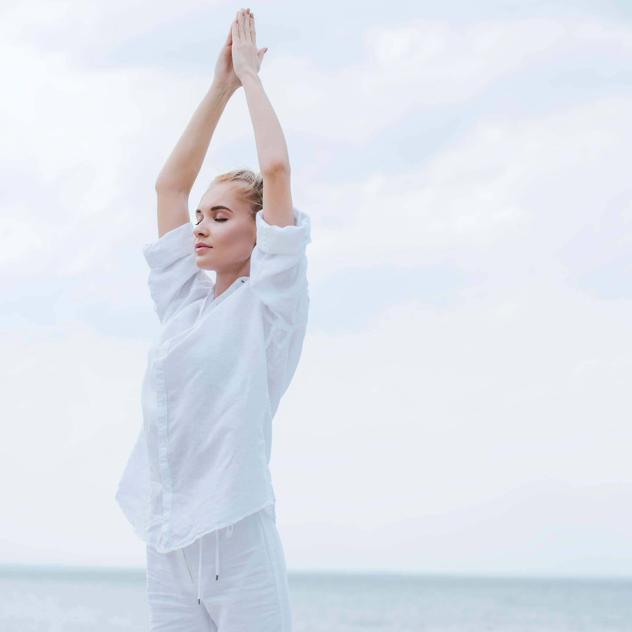 Yogaübung im Freien Eine Frau in heller Kleidung hält die Hände bei einer Yogaübung über dem Kopf zusammen.